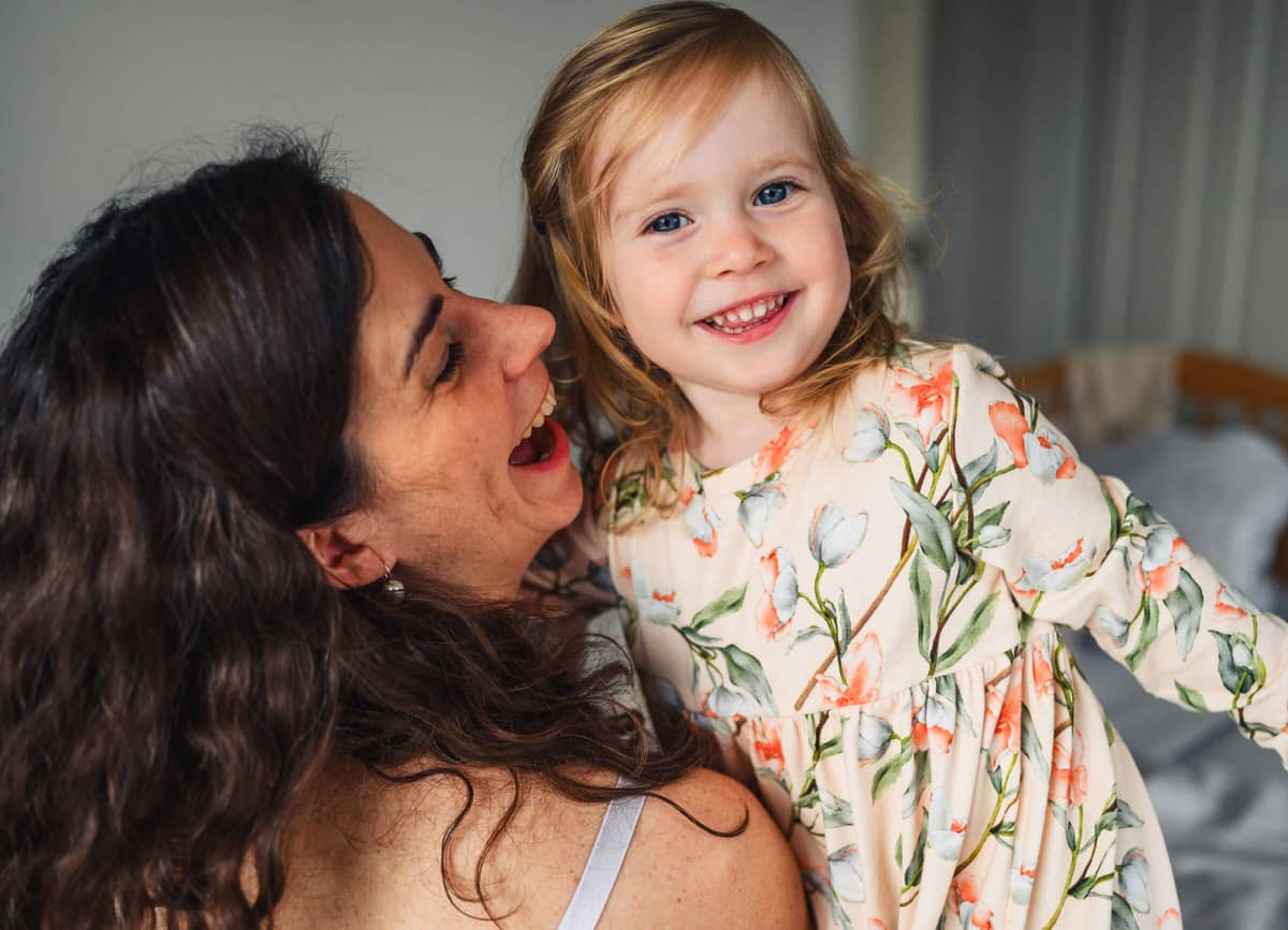 Mama und lachende Tochter im geblümten Kleid kuscheln bei einer Homestory zu Hause, die Tochter schaut strahlend in die Kamera