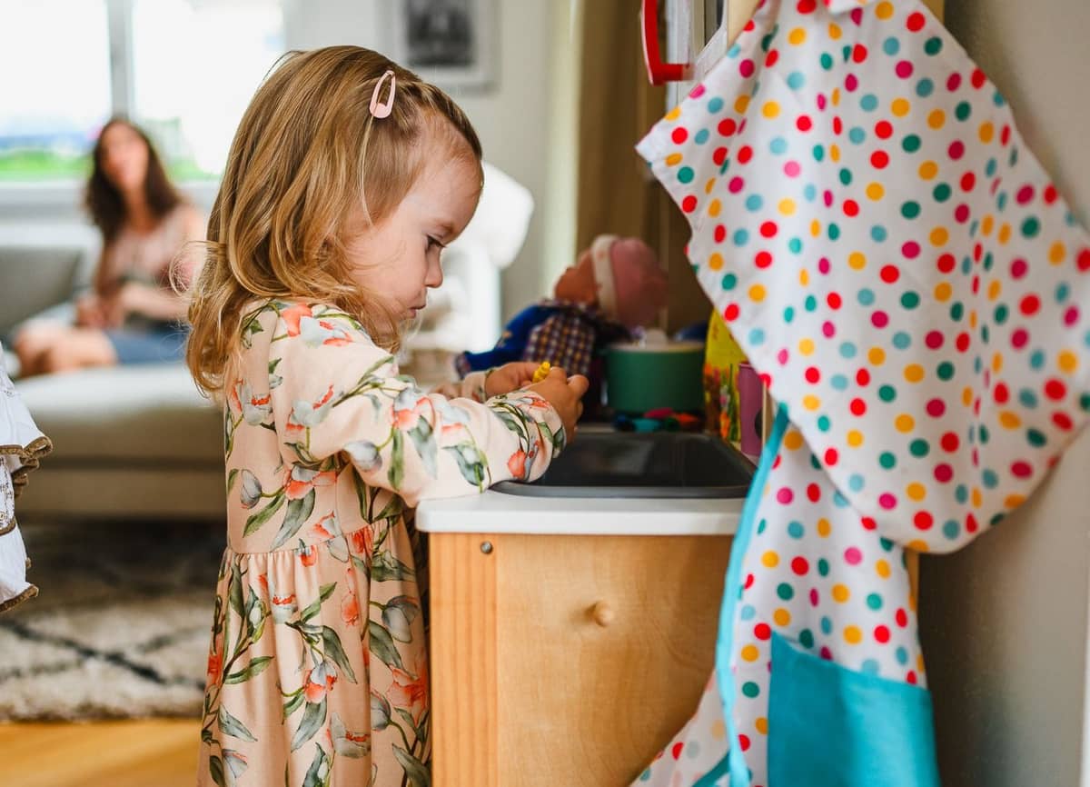 Kleines Mädchen spielt im Kinderzimmer, im Hintergrund Mama mit Baby