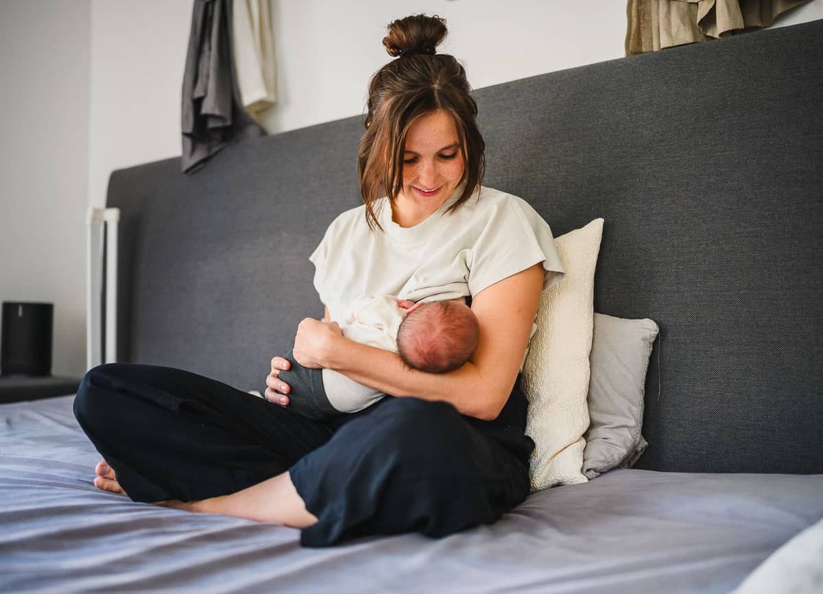 Mama sitzt im Schneidersitz auf dem Bett und hält ihr Baby im Arm