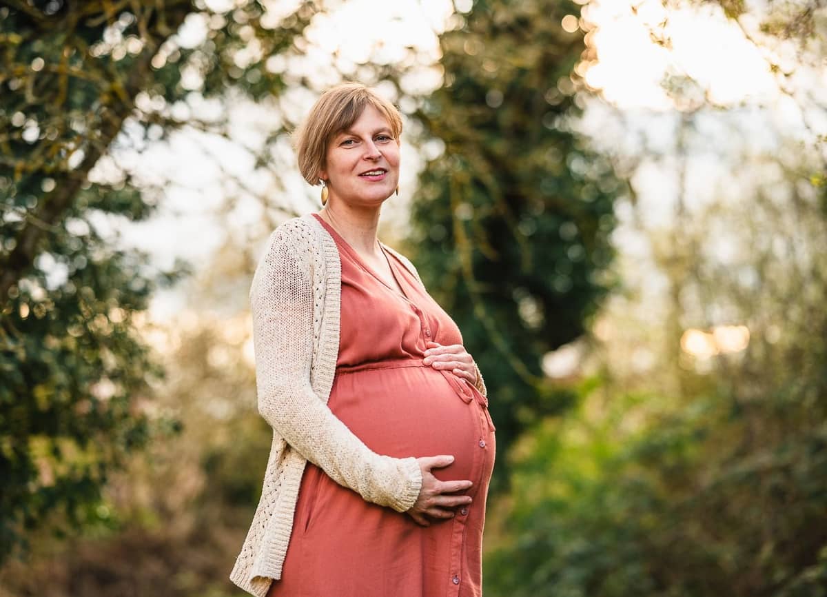 Schwangere Frau im roten Kleid und heller Strickjacke hält ihren Babybauch