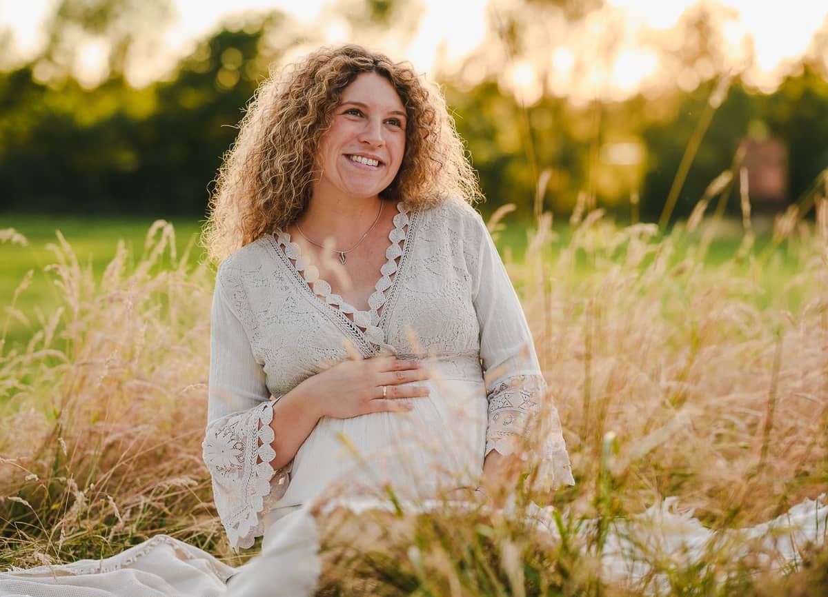 Schwangere Frau sitzt lachend im Gras mit den Händen am Babybauch