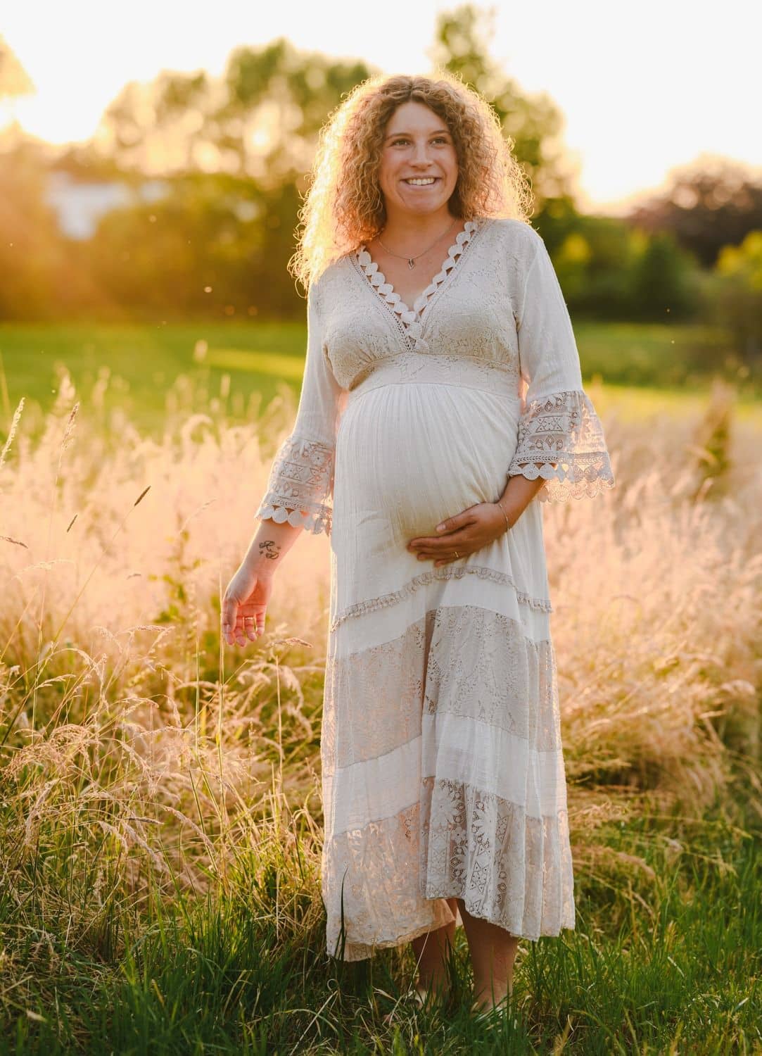 Schwangere Frau im weißen Kleid steht im goldenen Gras und hält ihren Babybauch