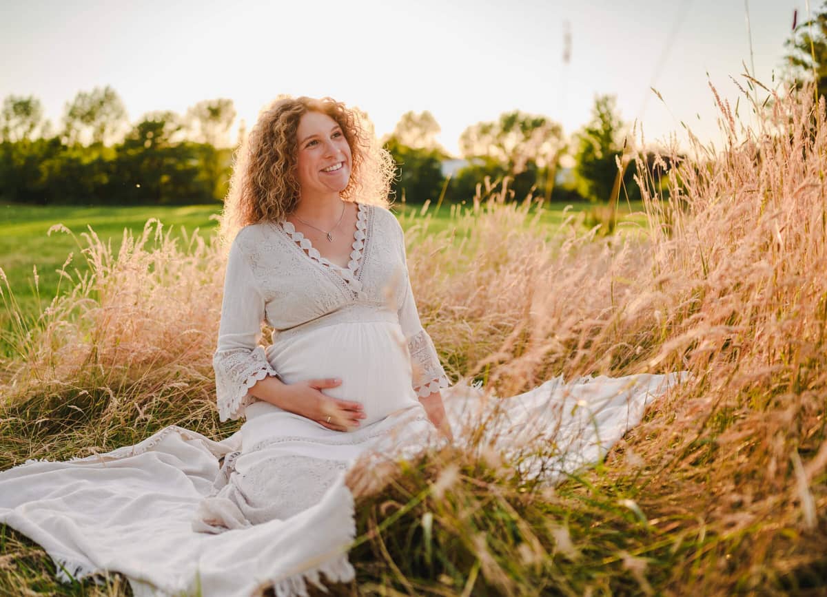 Schwangere Frau im weißen Spitzenkleid sitzt im hohen Gras und hält lächelnd ihren Babybauch im Abendlicht