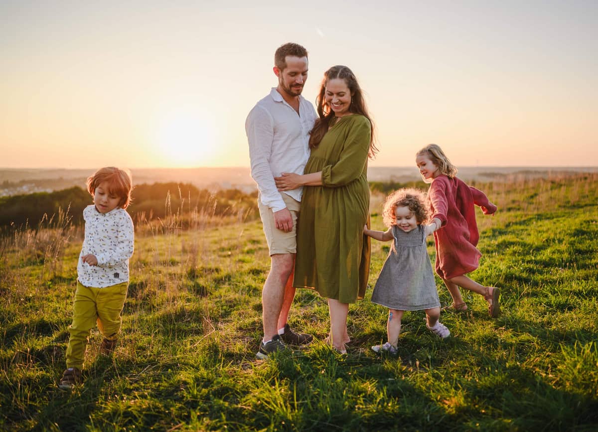 Familie mit drei Kindern auf einer Wiese bei Sonnenuntergang, die Eltern halten sich im Arm, die Kinder tanzen um sie herum