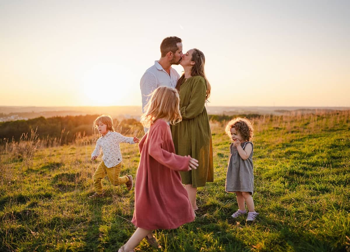 Familie auf einer Wiese bei Sonnenuntergang, die Eltern küssen sich, die Kinder spielen