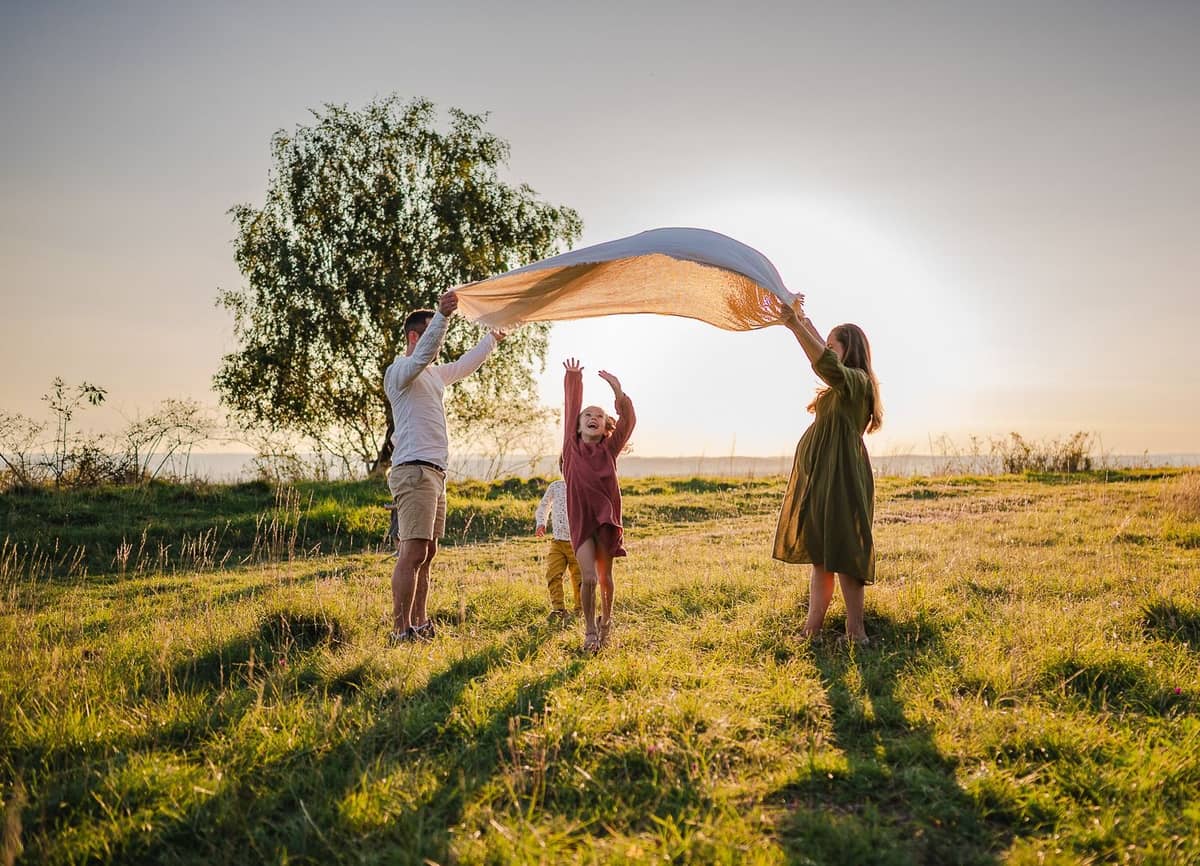 Familie lässt ein großes Tuch im Wind wehen, im Hintergrund ein Baum